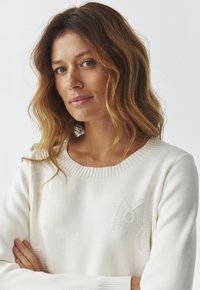 Suéter blanco de punto con un patrón texturizado en el frente y un cuello de canalé. La modelo tiene el cabello ondulado y está cruzando los brazos.