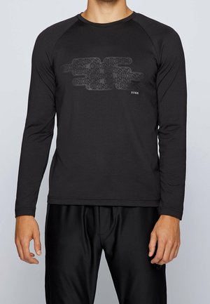 Man draagt een zwart shirt met lange mouwen met herhaalde "BOSS"-tekst in donkergrijs aan de voorkant en bijpassende zwarte broek tegen een lichtgrijze achtergrond.