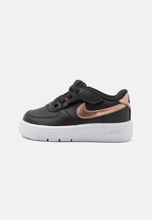 Baskets pour tout-petit noires avec le logo Nike en rose doré, lacets et fermeture Velcro, et semelle blanche sur fond gris clair.