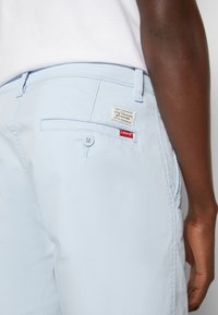 Gros plan sur une personne portant un short Levi's bleu clair, montrant la poche arrière avec un bouton et les étiquettes de la marque Levi's.