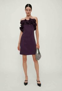 Μωβ φόρεμα με ρυτιδωτό ντεκολτέ, εφαρμοστή σχεδίαση και μήκος πάνω από το γόνατο. Συνδυάζεται με μια μικρή υφασμάτινη τσάντα και μαύρες γόβες.