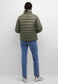 Olijfgroene, gewatteerde pufferjack met hoge kraag; gecombineerd met blauwe jeans en witte sneakers. Vloeiende textuur, modern ontwerp.