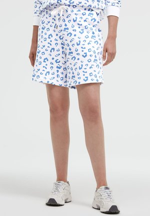 Person trägt weiße Shorts mit blauem Leopardenmuster, weißem Kordelzug und weißen Sneakers, steht vor einem einfarbigen Hintergrund.