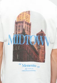 Visoke mestne zgradbe, osvetljene z toplo svetlobo, z napisom "MIDTOWN" v velikih modrih črkah in besedilom "Memento NY 10036 Originals Studio" spodaj.