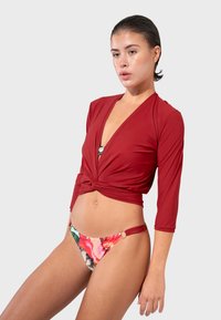 Crvena bluzica s tričetrt rukavima i čvorom na struku, u kombinaciji s donjim dijelom bikinija u više boja s cvjetnim uzorkom i bočnim dekoracijama.