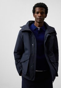 Marinblå vattentät jacka med dragkedja framtill, snodd i huvan och sidofickor. Bärs över en mörkblå polo t-shirt. Slät textur, modern design.