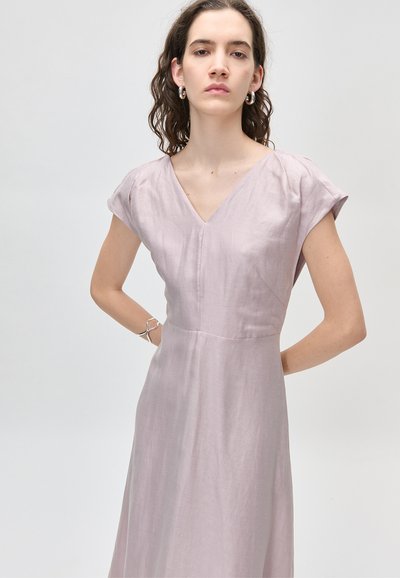 Νεαρή γυναίκα με σγουρά μαλλιά που φοράει ασημένια σκουλαρίκια κρίκους, ένα ανοιχτό μοβ φόρεμα με V λαιμόκοψη και ένα κυματιστό ασημένιο βραχιόλι στο αριστερό καρπό.