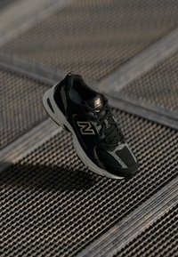 Sapatilha de corrida New Balance em preto e cinza sobre uma superfície de grelha de metal com padrões texturizados sob uma suave luz natural.