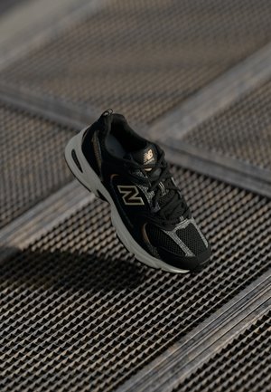 Baskets de course New Balance noires et grises sur une surface en grille métallique avec des motifs texturés sous une lumière naturelle douce.