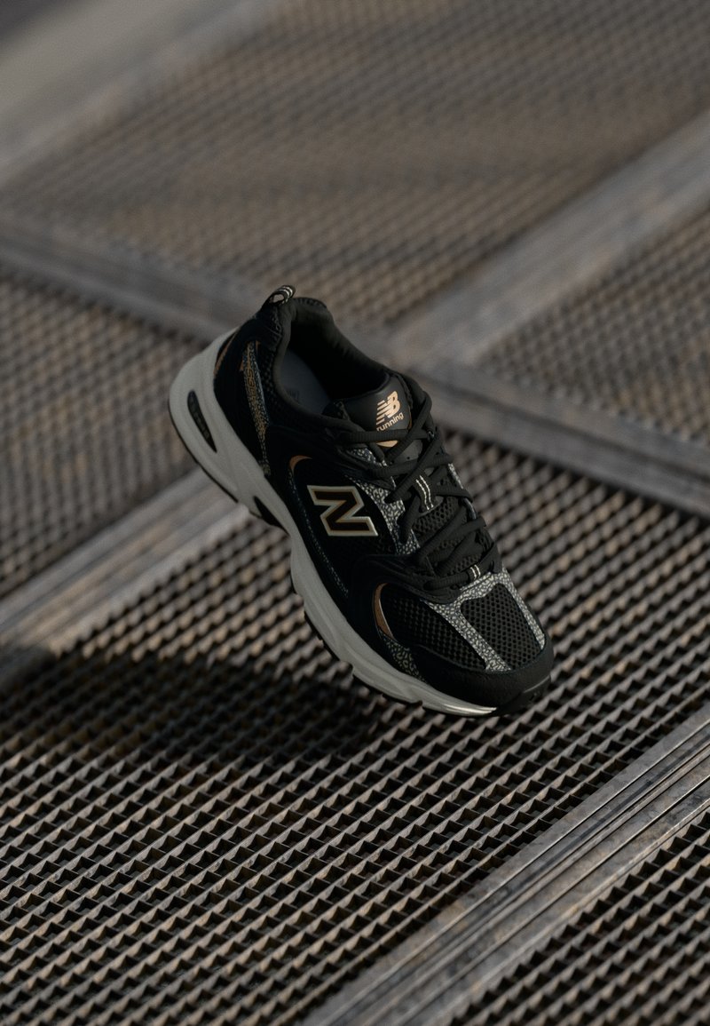 Sapatilha de corrida New Balance em preto e cinza sobre uma superfície de grelha de metal com padrões texturizados sob uma suave luz natural.
