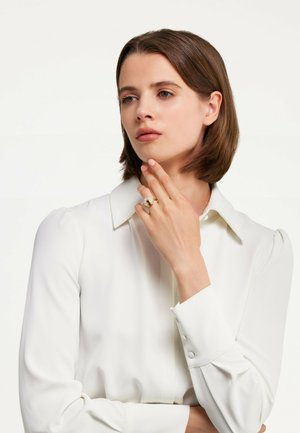 Eine Frau trägt eine weiße Bluse mit Kragen und Knopfdetails und hält ihre Hand nahe an einem goldenen Ring mit strukturierten Akzenten.
