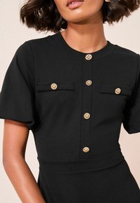 Rochie neagră cu mânecă scurtă, cu decolteu rotund, având două buzunare pe piept și detalii cu nasturi aurii pe față. Textura materialului este netedă.