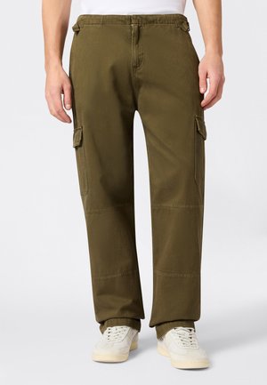 Person trägt olivgrüne Cargo-Hosen mit Seitentaschen und weiße Turnschuhe, steht vor einem schlichten, hellen Hintergrund.