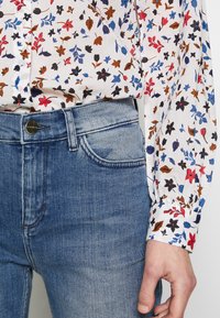 Chemise à manches longues florale avec des motifs rouges, bleus et bruns, associée à un jean en denim bleu clair avec une texture délavée et un bouton en métal.