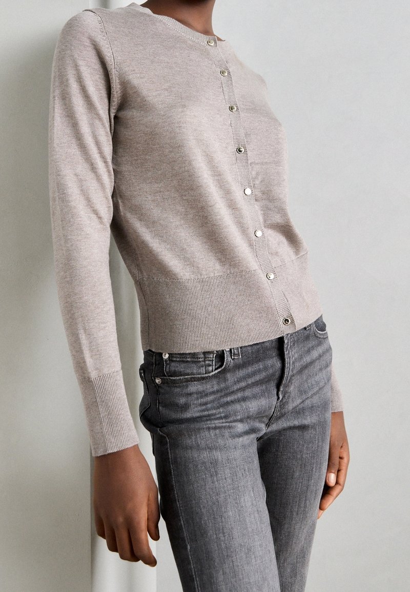Personne portant un cardigan beige boutonné à manches longues et un jean gris foncé ajusté sur un fond gris clair uni.