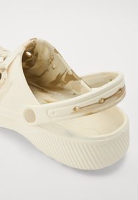 Beige and cream clog with textured insole, decorative holes, and stud accents on the strap. Made from flexible material, with a ridged sole.