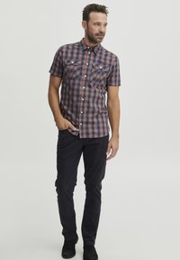 Camisa de cuadros de mangas cortas en tonos marino y óxido, con dos bolsillos en el pecho y diseño de botones, combinada con pantalones oscuros y botas negras.