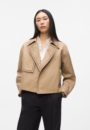 Mujer con cabello largo y negro, lleva una chaqueta corta beige sobre una camisa blanca y pantalones negros, de pie con las manos en los bolsillos.