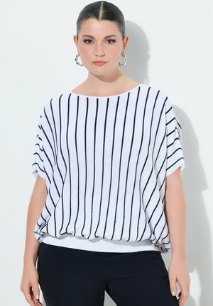 Mujer con el cabello peinado hacia atrás, lleva pendientes de aro plateados y una blusa blanca de manga corta con rayas verticales azul marino, combinada con pantalones oscuros.