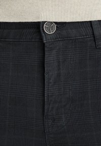 Mörkgrå rutiga byxor med en metallknapp graverad med "PME Legend Jeans" och en beige ribbad tröja instoppad.