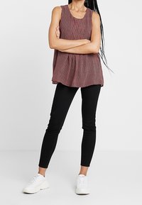 Top sin mangas con patrón rojo y blanco, combinado con pantalones ajustados negros y zapatillas blancas, de pie sobre un fondo liso.