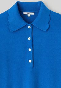 Polo bleu tricoté avec un col festonné, une patte de boutonnage à cinq boutons blancs et une texture lisse. L'étiquette de taille indique "S" de la marque BZB.