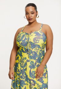 Robe jaune avec des motifs floraux paisley bleu et vert, dotée de bretelles réglables et d'un détail central froncé pour une silhouette ajustée.