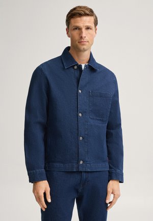Uomo che indossa una giacca di jeans blu scuro con bottoni frontali e tasca sul petto, abbinata a pantaloni di jeans blu scuro coordinati.