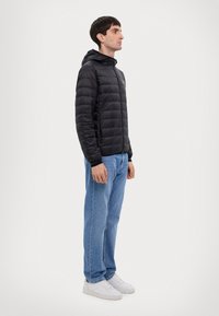 Giubbotto imbottito nero con cappuccio, dotato di una chiusura lampo frontale, polsini elastici e un design leggero, abbinato a jeans blu e sneakers bianche.