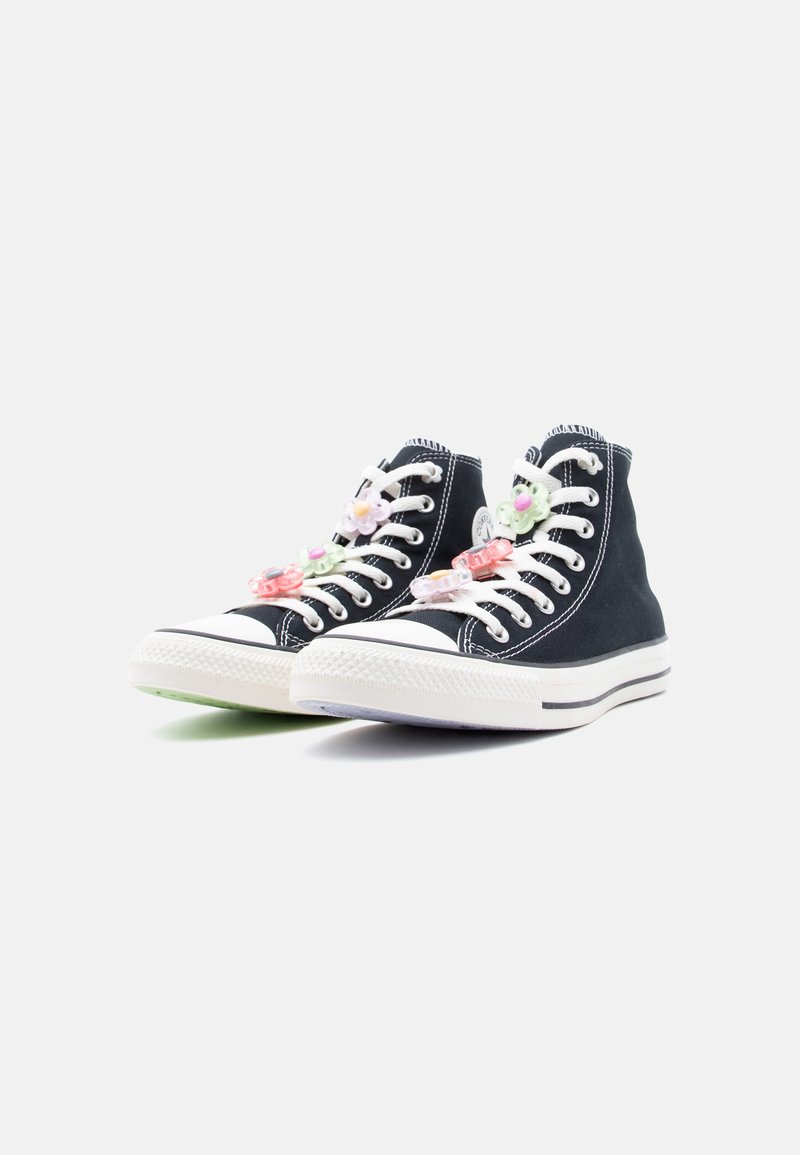 Sneakers alte nere con suole e lacci bianchi, decorate con ciondoli a forma di fiori di plastica color pastello sui lacci, su sfondo bianco.