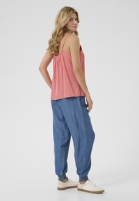 Roza brezrokavnik z čipkastim detajlom, v kombinaciji z ohlapnimi modrimi joggerskimi hlačami z elastičnimi manšetami in belimi športnimi čevlji.