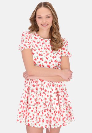 Fiatal nő hosszú, hullámos hajjal, mosolyogva, fehér, rövid ujjú ruhát visel, piros cseresznye mintával és keresztezett karokkal áll.