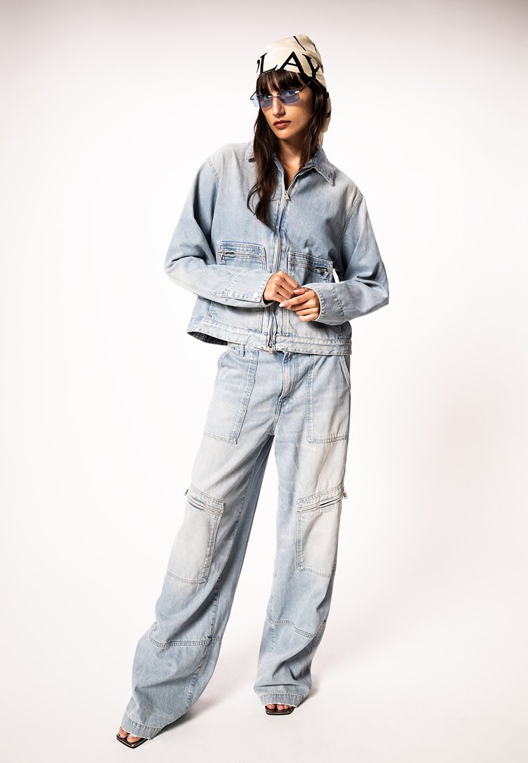 Γυναίκα που φοράει ανοιχτό μπλε υπερμεγέθη τζιν μπουφάν και παντελόνι, γυαλιά ηλίου με μπλε απόχρωση, μπεζ μαντίλι κεφαλής με μαύρο κείμενο και μαύρες ανοιχτές γόβες.