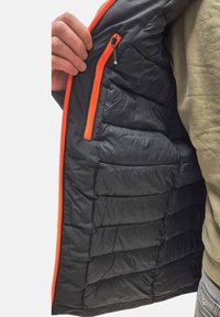 Giacca a vento nera con design trapuntato, dotata di zip arancione e dettagli delle tasche. L'interno presenta una superficie liscia e lucida.