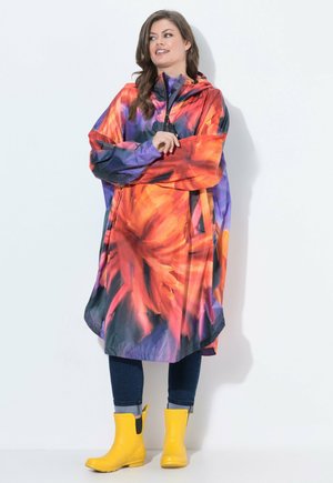 Mujer con un impermeable colorido naranja y púrpura con capucha y botas de lluvia amarillo brillante, de pie frente a un fondo blanco.