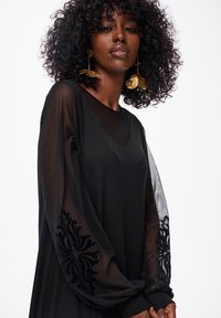 Frau mit lockigem Haar, die große goldene Blumenohrringe und ein schwarzes Kleid mit durchsichtigen bestickten Ärmeln trägt, vor einem weißen Hintergrund steht.