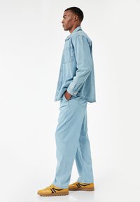 Lichtblauwe denim blouse met een zak, gecombineerd met bijpassende lichtblauwe broeken. De schoenen zijn gele met zwarte accenten en textuurlijke zolen.