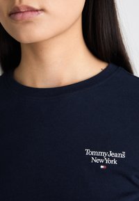 Tumed sinine puuvillane T-särk ümmarguse kaeluse ja lühikeste varrukatega, millel on valge kantud logo: "Tommy Jeans New York" ja värviline aktsent.