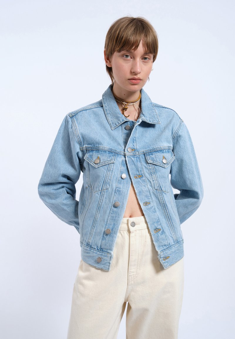 Chaqueta de mezclilla azul claro con cuello tipo solapa, bolsillos frontales y botones plateados. Presenta un corte relajado y una textura ligeramente desgastada.
