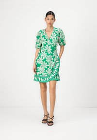 Groene bloemenjurk met pofmouwen, een wikkelontwerp en geborduurde details. Gecombineerd met zwarte sandaaltjes met banden.