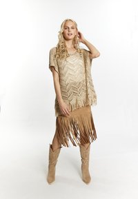 Poncho beige de ganchillo con patrón de chevrón y detalles de flecos, combinado con una falda de flecos color marrón claro y botas de cowboy marrón claras con punta. 