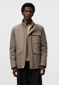 Giacca marrone imbottita con colletto alto, tasche frontali, bottoni a pressione e chiusura con zip, indossata sopra un maglione leggero.