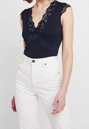 Femme portant un haut noir sans manches avec des détails en dentelle et un pantalon blanc taille haute, debout devant un fond clair uni.