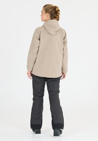 Beige vedenkestävä takki hupulla, yhdistetty mustiin eristyspolkimiin. Takissa on suora leikkaus ja housuissa on vetoketjulliset sivuventtiilit.