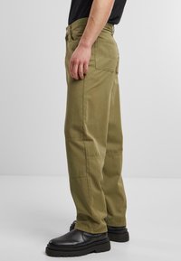 Pantalones cargo de color verde oliva hechos de algodón, con un ajuste relajado, bolsillos laterales y detalles de costura. Combinados con zapatos negros texturizados.
