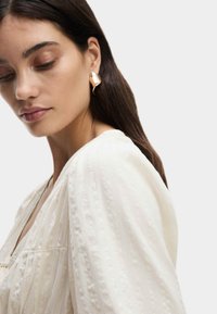 Blusa fluida color crema con strisce testurizzate; scollatura rotonda e maniche a sbuffo. Orecchini astratti in tono oro completano l'outfit.