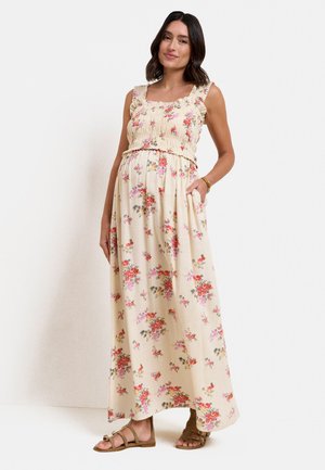 Frau trägt ein ärmelloses cremefarbenes Maxikleid mit pinkem und rotem Blumenmuster, steht mit einer Hand in der Tasche und braunen Sandalen.