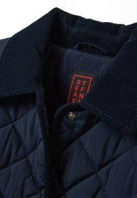 Giacca trapuntata blu navy con colletto in velluto, chiusura a scatto e etichetta rossa del marchio. Caratterizzata da un distintivo motivo a rombi su tutta la superficie.