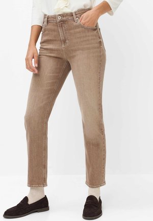 Persoon die lichtbruine strechtbroeken, witte sokken, donkerbruine loafers en een wit shirt met lange mouwen draagt, met één hand in de zak.
