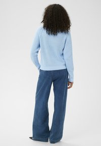 Personne avec des cheveux bouclés portant un pull en tricot bleu clair et un jean bleu foncé à large jambe, se tenant de dos.